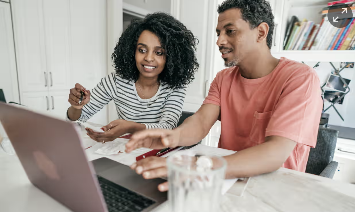 a husband and wife look at a laptop together as they plan their business